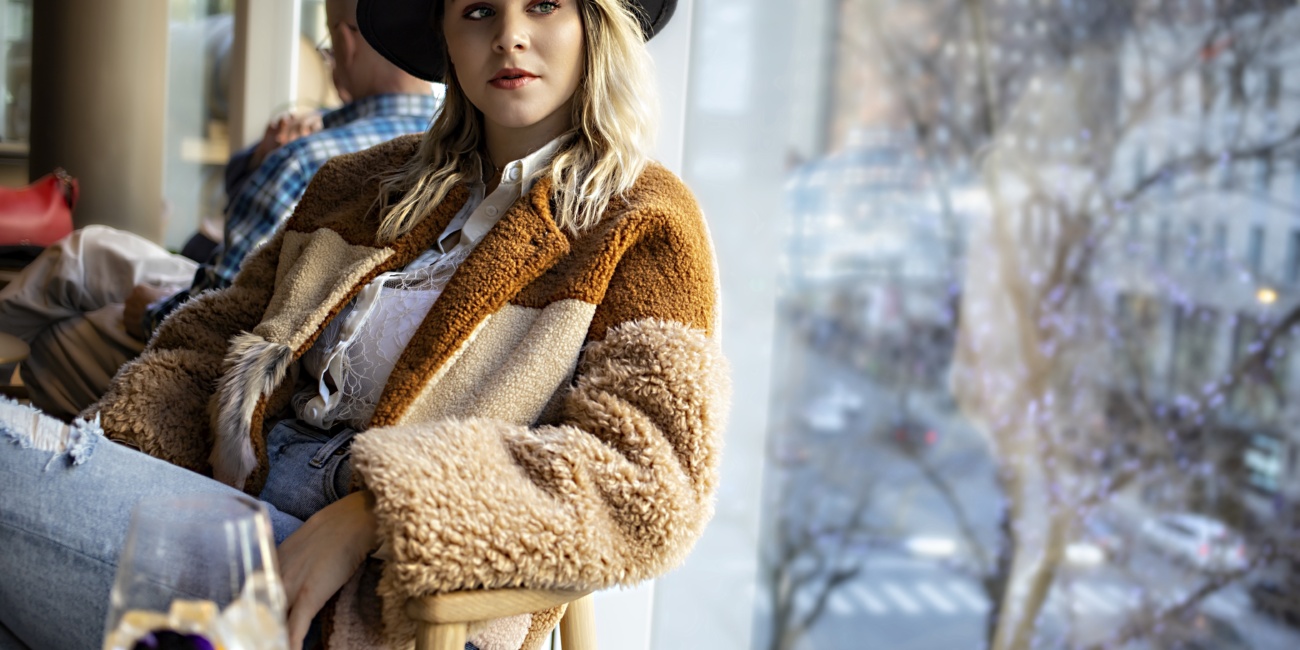 A young model in a cozy sherpa jacket and wide-brimmed hat sits by a café window, gazing thoughtfully outside - capturing the reflective mood of balancing runway glamour with the emotional challenges of the holiday season.